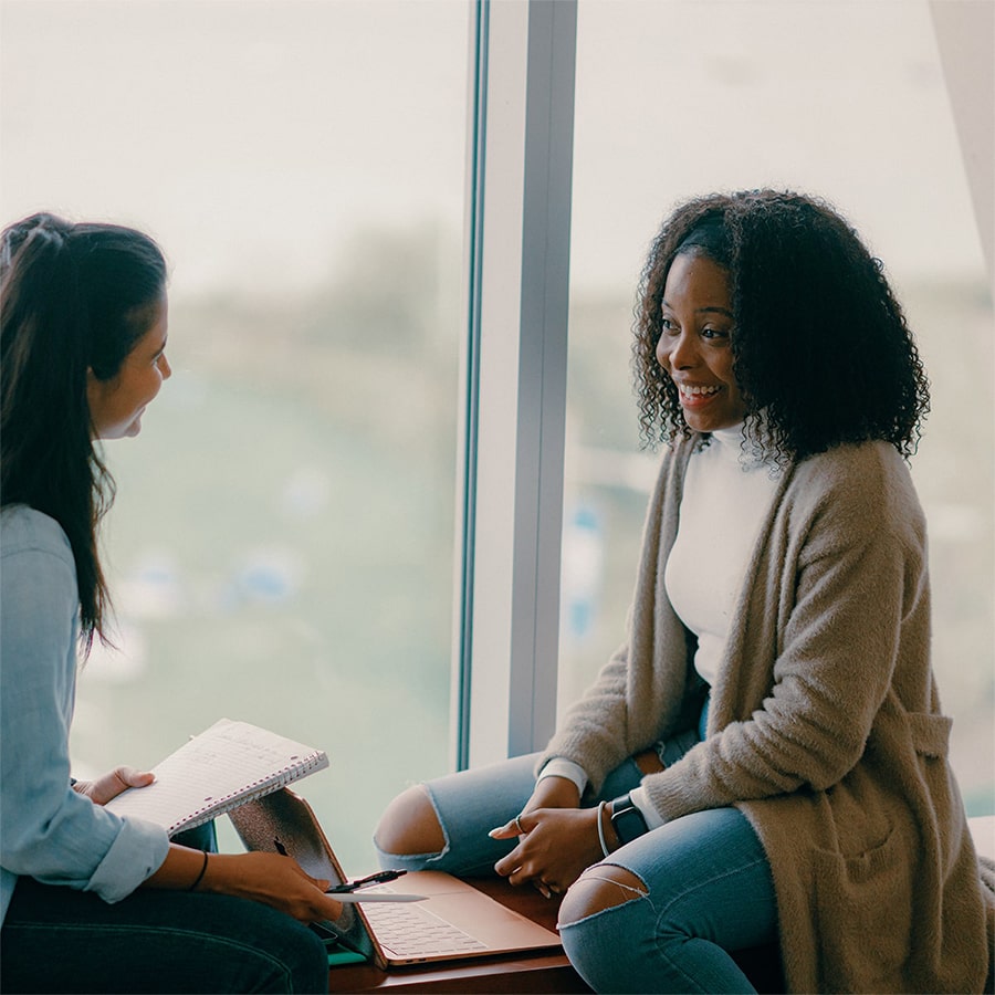Two female students talking and working on their notebooks and laptop in front of large window.