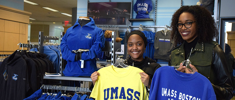Two smiling women holding up UMass Boston apparel in a campus bookstore, surrounded by racks of university-branded clothing including sweatshirts and jackets.