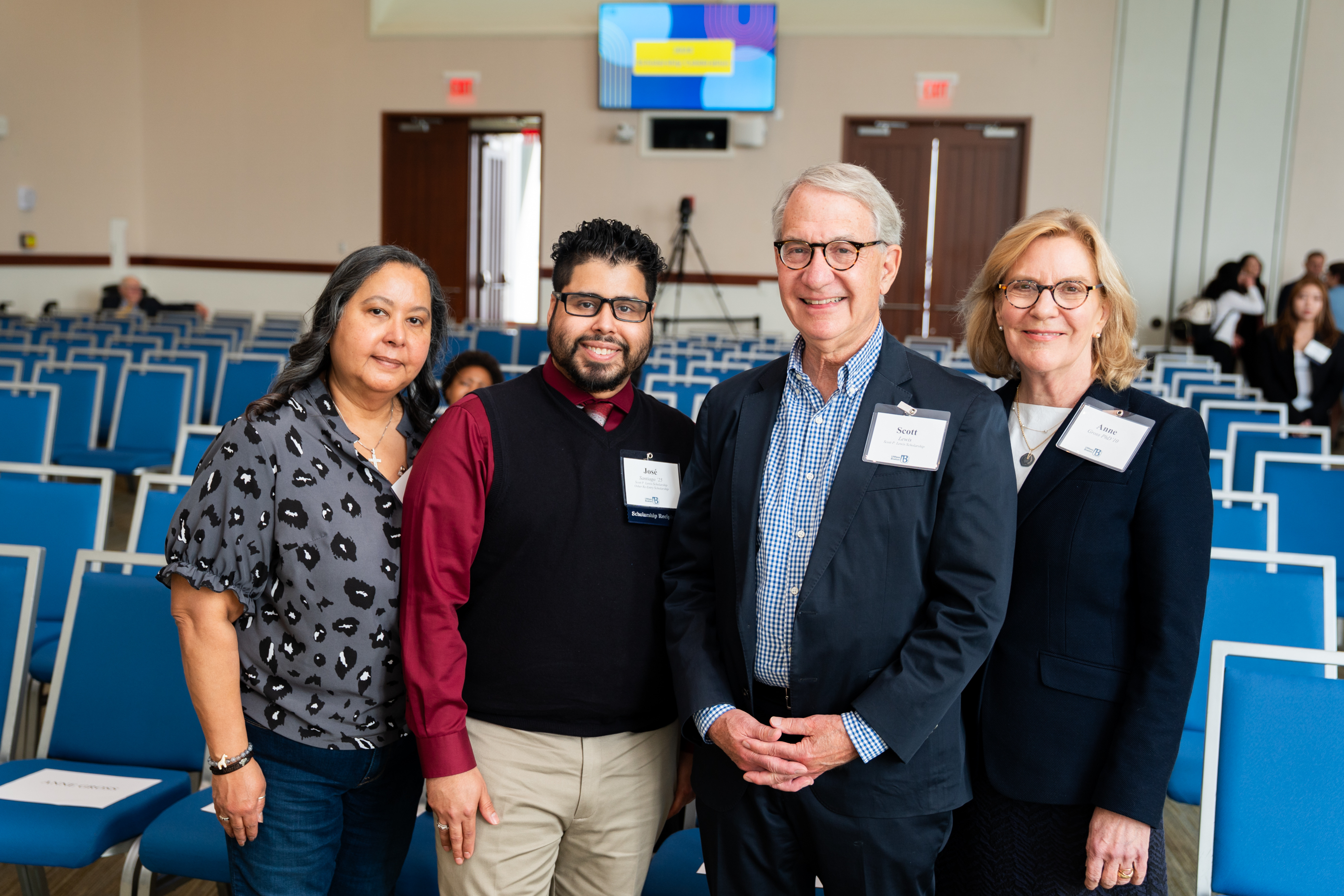 Four individuals, Jose and his mother, Scott Lewis, and Anne Gross, smile for a photo in a bright auditorium with blue chairs.