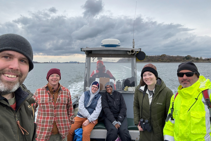 Six people bundled in winter coats and hats pose on a small boat during a trip to the Boston Harbor Islands, with gray clouds overhead and the city skyline in the distance.
