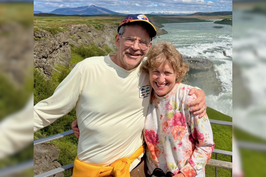 A smiling couple poses together at an overlook with a large waterfall and mountains in the background.
