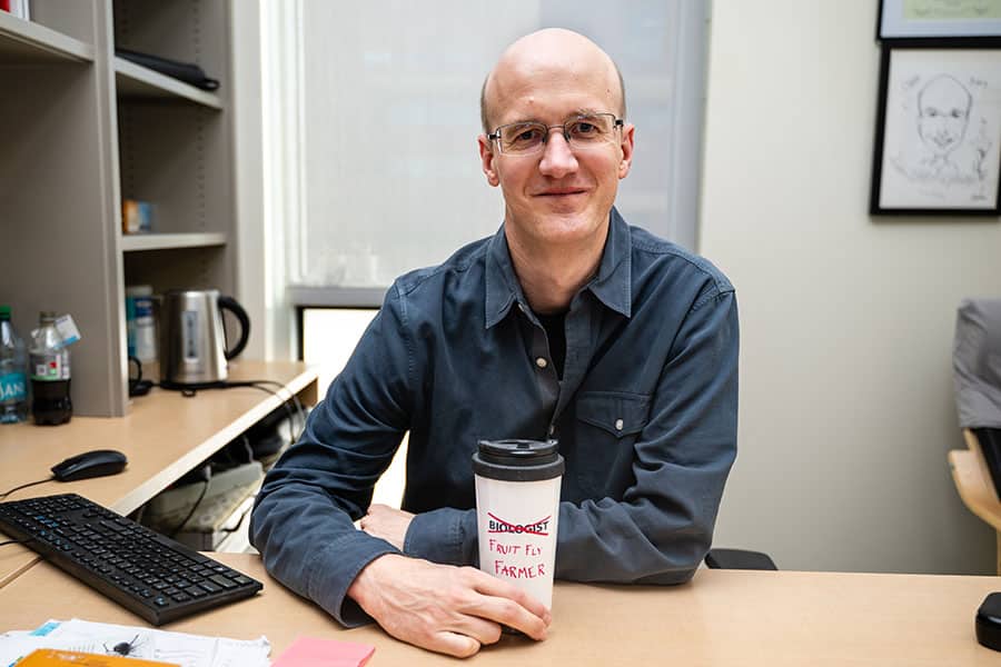 Jens Rister sits at his desk, smiling at the camera. He holds a cup with the word a 