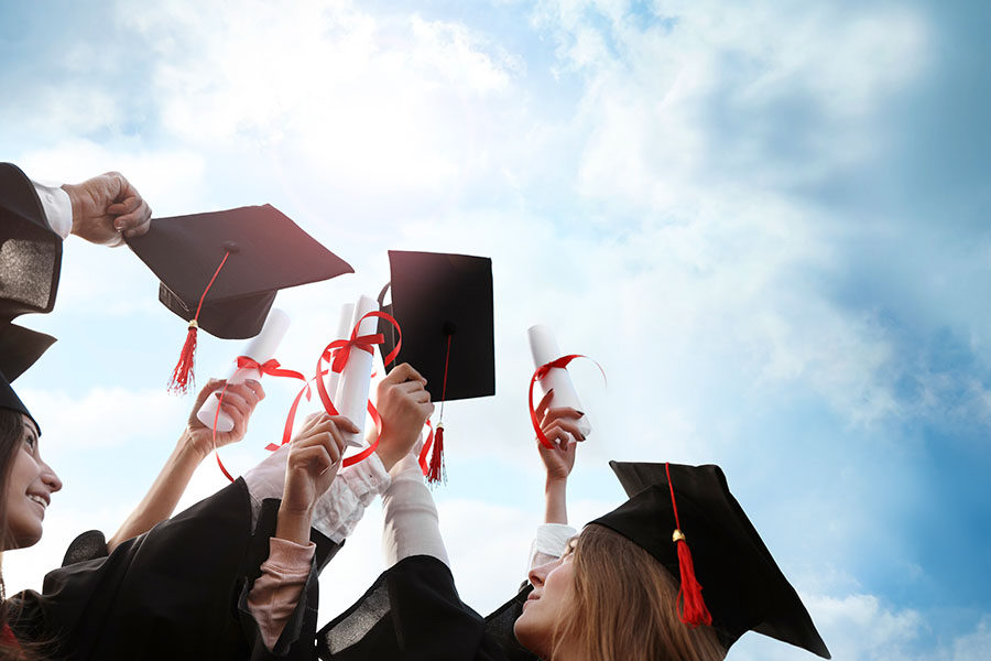 Group of graduates in black caps and gowns raising diplomas and mortarboards into a bright blue sky, celebrating their achievement.