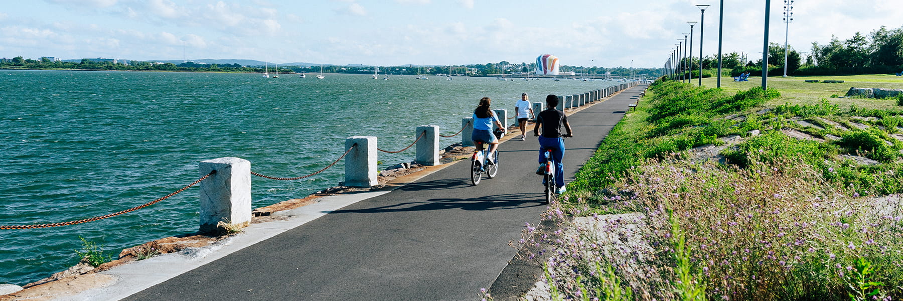 Two people bike on the Harborwalk going toward the gas tank with Corita Kent mural.
